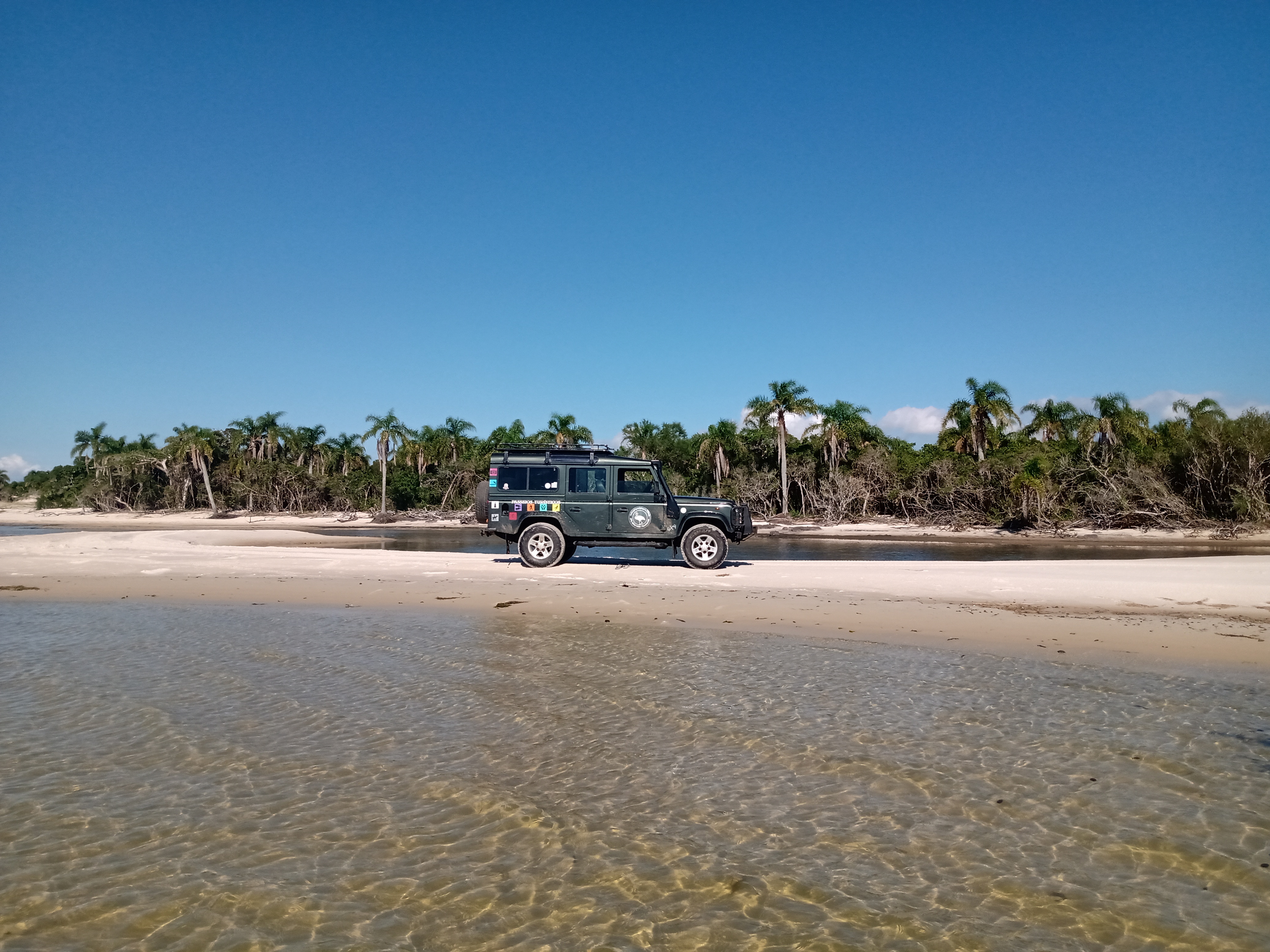 beira da lagoa com veículo na areia e coqueiros ao fundo