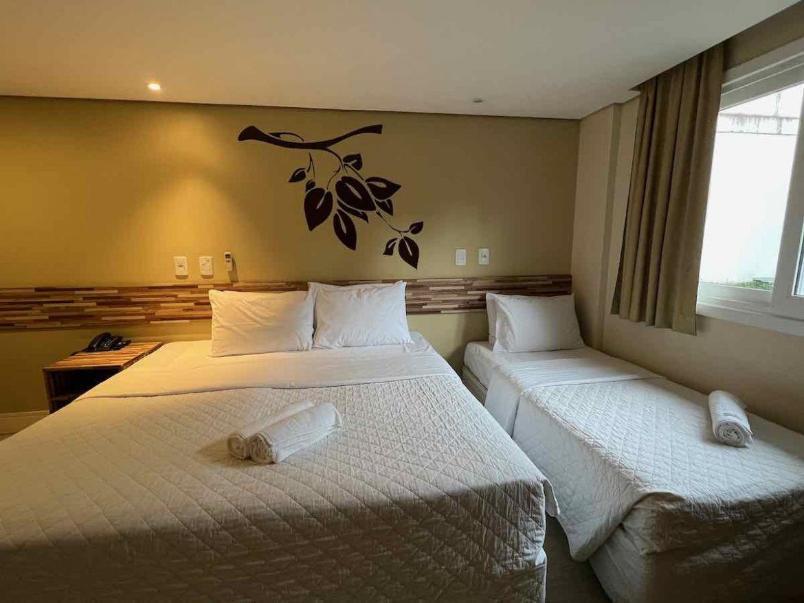 O que fazer em Novo Hamburgo: o quarto do hotel tem cama de casal e solteiro com roupa de cama branca