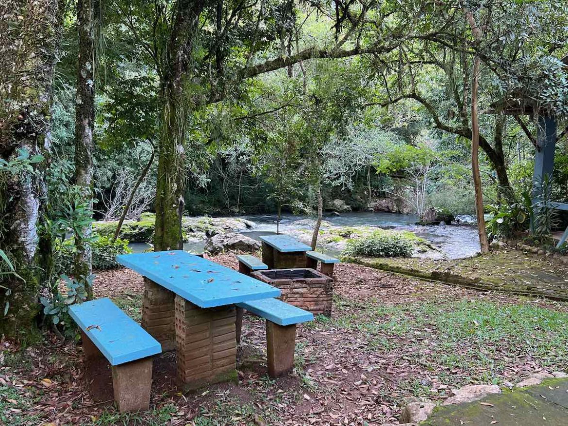 Sapiranga (RS): na foto há muitas árvores e no canto esquerdo uma mesa de madeira com bancada azul, próximo a um riacho